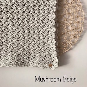 May include: A crocheted blanket in a mushroom beige color. The blanket features a detailed shell stitch pattern and a scalloped edge. The text "Mushroom Beige" is displayed at the bottom of the image. A wooden tag is attached to the blanket.