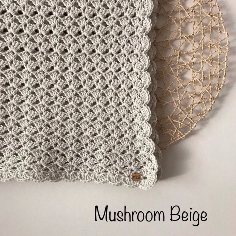 May include: A crocheted blanket in a mushroom beige color. The blanket features a detailed shell stitch pattern and a scalloped edge. The text "Mushroom Beige" is displayed at the bottom of the image. A wooden tag is attached to the blanket.