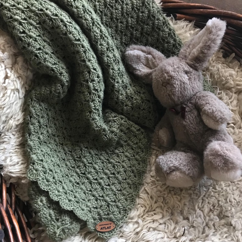 May include: A sage green crocheted blanket with a scalloped edge and a plush gray bunny toy. The blanket has a textured pattern and a small wooden tag that reads "handmade by ATLAS". The items are on a white, fluffy surface.