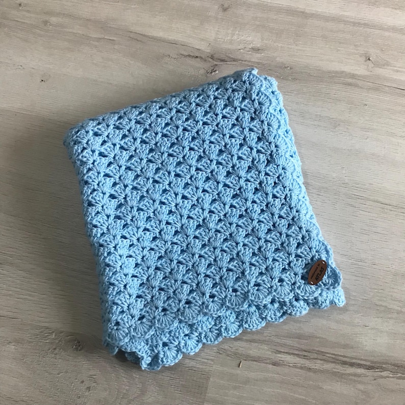 May include: A light blue, handmade crochet blanket with a scalloped edge. The blanket features a detailed openwork pattern and a small, round, brown tag. The blanket is folded and lies on a light wood surface.