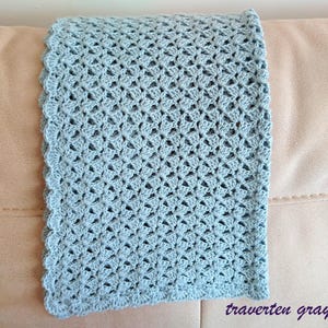 May include: A crocheted blanket in a soft, muted blue-gray color. The blanket features a delicate openwork pattern and a scalloped edge. The text "traverten gray" is visible in the bottom right corner.