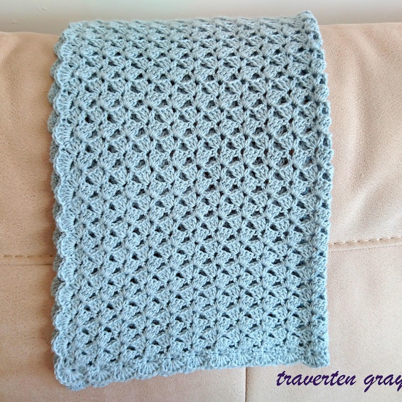 May include: A crocheted blanket in a soft, muted blue-gray color. The blanket features a delicate openwork pattern and a scalloped edge. The text "traverten gray" is visible in the bottom right corner.