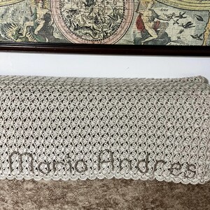 May include: A light gray crocheted blanket with the name "Maria Andres" embroidered in a matching thread. The blanket features a detailed openwork pattern and a scalloped edge. A vintage map is in the background.