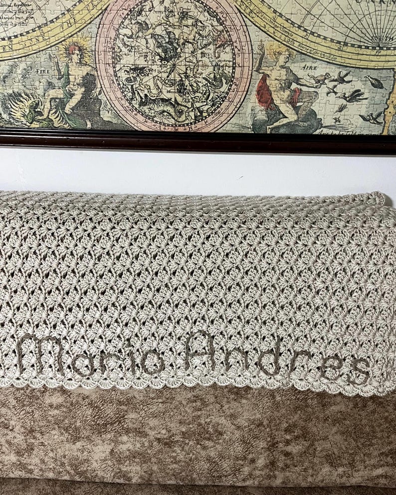 May include: A light gray crocheted blanket with the name "Maria Andres" embroidered in a matching thread. The blanket features a detailed openwork pattern and a scalloped edge. A vintage map is in the background.