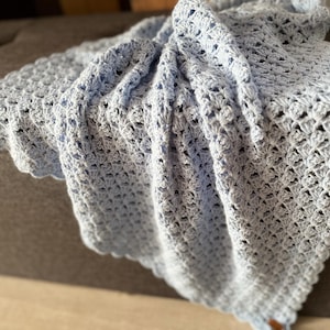 May include: A light blue crocheted blanket with a scalloped edge. The blanket has a delicate, open weave pattern, and is draped over a surface. The soft, pastel color and intricate design suggest a handmade item, suitable for a baby or as a decorative throw.