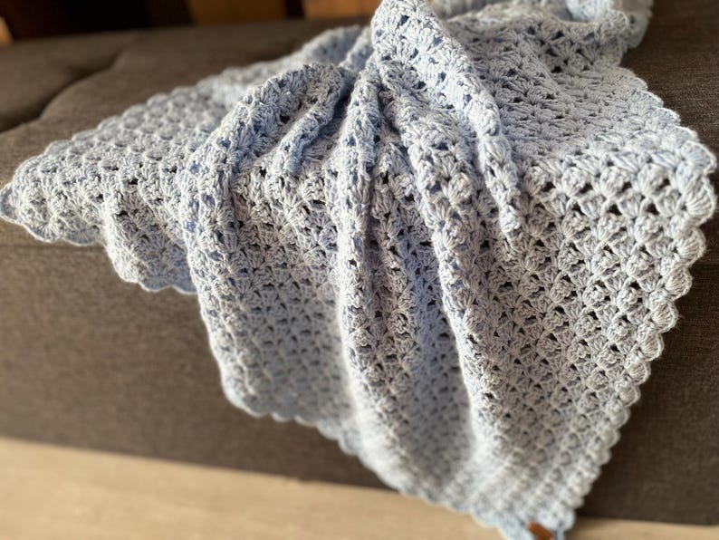 May include: A light blue crocheted blanket with a scalloped edge. The blanket has a delicate, open weave pattern, and is draped over a surface. The soft, pastel color and intricate design suggest a handmade item, suitable for a baby or as a decorative throw.