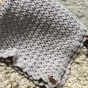 May include: A handmade, light gray crochet blanket with a scalloped edge. The blanket has a textured pattern and a small, brown leather tag that reads "Handmade by ATLAS". The blanket is folded on a textured, off-white surface.