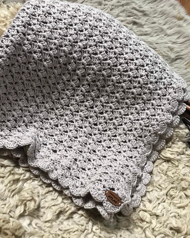 May include: A handmade, light gray crochet blanket with a scalloped edge. The blanket has a textured pattern and a small, brown leather tag that reads "Handmade by ATLAS". The blanket is folded on a textured, off-white surface.