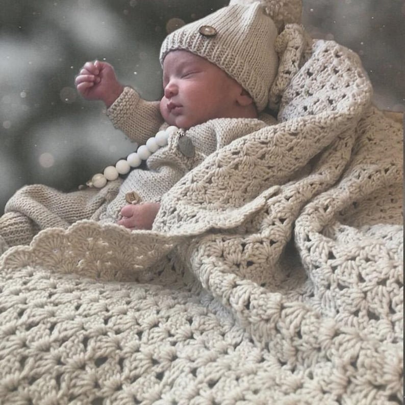 May include: A sleeping baby wrapped in a cream-colored crocheted blanket and matching hat. The baby is wearing a cream-colored sweater with wooden button details and a white beaded necklace. The blanket has a scalloped edge.