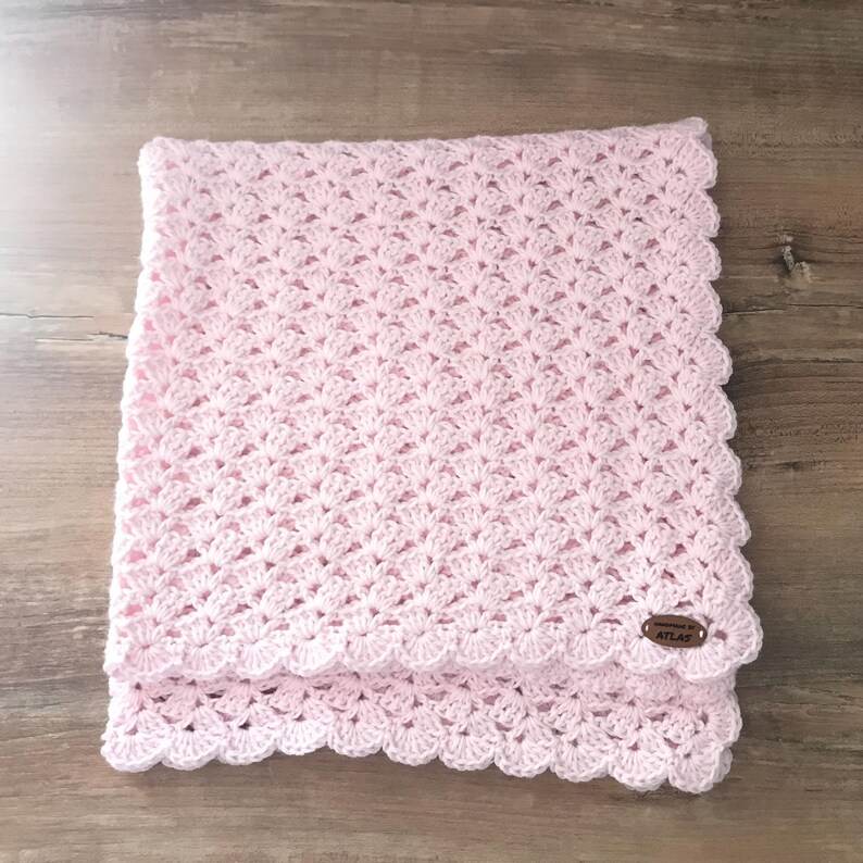 May include: A light pink crocheted blanket with a scalloped edge. The blanket is folded and has a small brown tag with the word "ATLAS" on it. The blanket is made of soft yarn and is likely for a baby.
