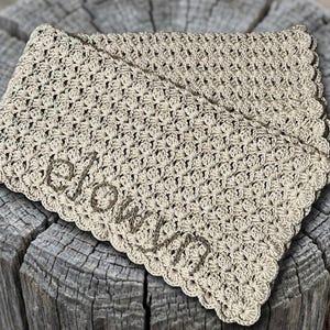 May include: A beige crocheted blanket with the name "elovvn" embroidered in a darker beige thread. The blanket has a scalloped edge and is folded over. The blanket is resting on a weathered wooden surface.