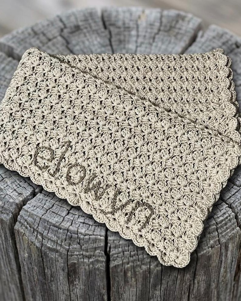 May include: A beige crocheted blanket with the name "elovvn" embroidered in a darker beige thread. The blanket has a scalloped edge and is folded over. The blanket is resting on a weathered wooden surface.