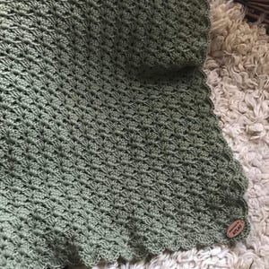 May include: A sage green crocheted blanket with a scalloped edge. The blanket has a textured, shell-like pattern. A small, round, wooden tag with the word "ATLAS" is attached to the blanket.