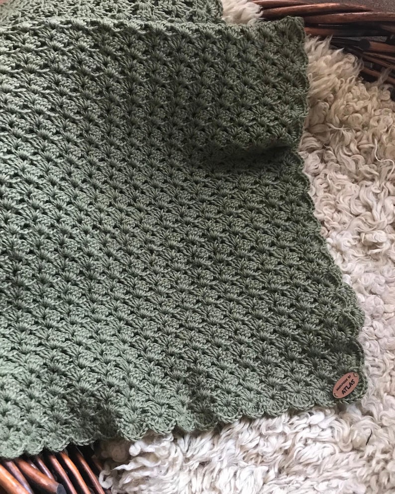 May include: A sage green crocheted blanket with a scalloped edge. The blanket has a textured, shell-like pattern. A small, round, wooden tag with the word "ATLAS" is attached to the blanket.