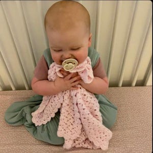 May include: A baby wearing a pink and green outfit, holding a light pink crocheted blanket. The baby is also wearing a pacifier. The blanket has a scalloped edge.
