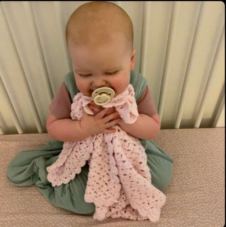 May include: A baby wearing a pink and green outfit, holding a light pink crocheted blanket. The baby is also wearing a pacifier. The blanket has a scalloped edge.