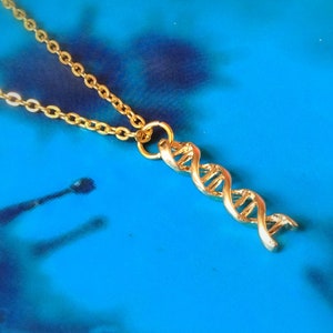 Può includere: Una collana dorata con un ciondolo a doppia elica del DNA. Il ciondolo è finemente dettagliato, mostrando la struttura a spirale. La catena e il ciondolo sono di un colore oro brillante, su sfondo blu. Un gioiello a tema scientifico.