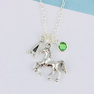 May include: Silver necklace with a horse pendant, a letter "A" charm, and a small green gemstone. The necklace is suspended from a delicate chain. The background is white and light blue.