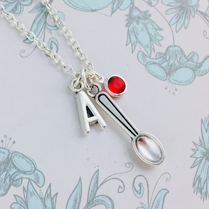Könnte beinhalten: Eine silberne Halskette mit einem Löffel-Anhänger, einem Buchstaben 'A'-Anhänger und einem roten Edelstein-Anhänger. Die Halskette liegt auf einem weißen Hintergrund mit einem Blumenmuster.