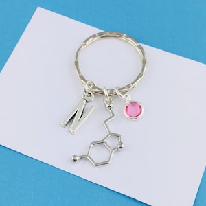 May include: Silver keychain with a pink crystal charm and a silver charm shaped like the chemical structure of dopamine. The keychain also has a silver charm with the letter "N" on it.