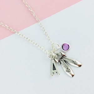 Puede incluir: Un collar de plata con un dije de zapatillas de ballet, un dije con la letra 'A' y una piedra preciosa púrpura.