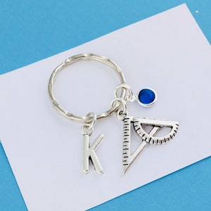 Puede incluir: Llavero plateado con un dije de letra "K", un dije de regla y transportador, y una gema azul. El llavero está sobre una tarjeta blanca sobre un fondo azul. Los dijes son detallados y brillantes.