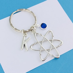 Op de afbeelding: Een zilverkleurige metalen sleutelhanger met een textuurrijke ronde ring, een letter K-bedel, een zilveren atoom-symbool met kruisende elliptische banen en een kleine ronde bedel met een diepblauwe steen.