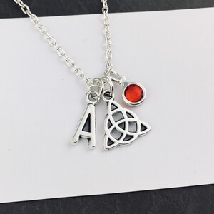 Peut inclure: Un collier de couleur argentée avec trois breloques : une lettre majuscule A, un symbole de nœud triquetra et une petite breloque circulaire avec une pierre précieuse rouge vif. Les breloques sont suspendues à une fine chaîne argentée.