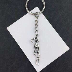 Könnte beinhalten: Ein silberfarbener Metallschlüsselanhänger mit einem Skelett-Anhänger, der an einem runden Spaltring befestigt ist. Die Skelettfigur ist in stehender Pose mit einem hinter den Kopf erhobenen Arm dargestellt und weist detaillierte Rippen und Gliedmaßen auf.