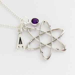 May include: A silver-toned necklace featuring a metal atom pendant, a small charm shaped like the letter A, and a circular purple gemstone charm. The necklace is composed of a delicate silver-coloured chain that connects to the three hanging charms.