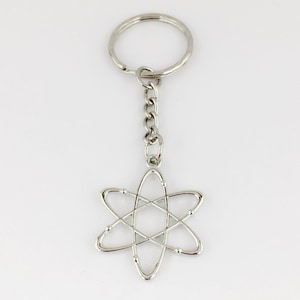 May include: A silver-coloured keyring with a chain and a pendant in the shape of an atom. The atom pendant is a stylized design. The keyring is made of metal and is set against a white background.