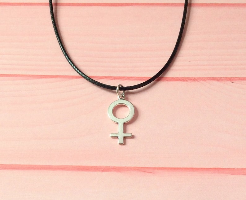 May include: A silver-colored pendant in the shape of the Venus symbol hangs from a black cord necklace. The necklace is displayed against a pink wooden background.