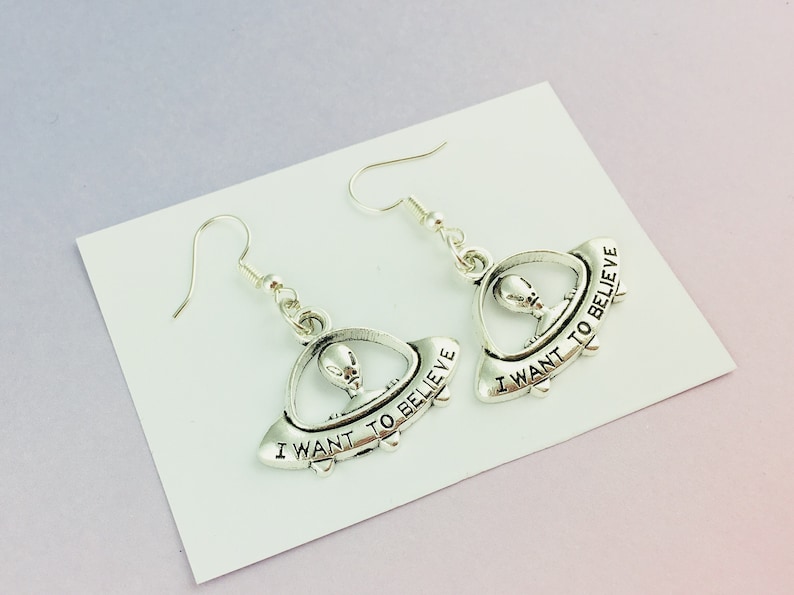 May include: Silver-toned alien earrings. Each earring features a small alien figure inside a UFO with the phrase "I WANT TO BELIEVE" inscribed on the side. The earrings have a hook closure.