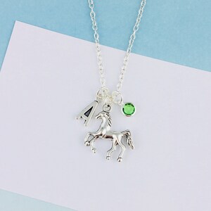 May include: A silver-toned necklace featuring a delicate chain with three charms: a silver letter A, a silver horse figurine in a prancing pose, and a small round green crystal bead. The charms are suspended from a single jump ring on the chain.