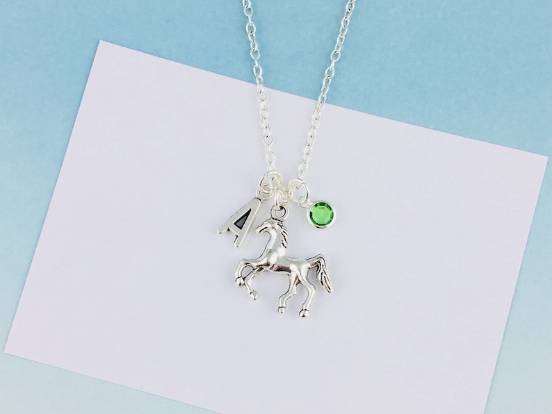 May include: A silver-toned necklace featuring a delicate chain with three charms: a silver letter A, a silver horse figurine in a prancing pose, and a small round green crystal bead. The charms are suspended from a single jump ring on the chain.