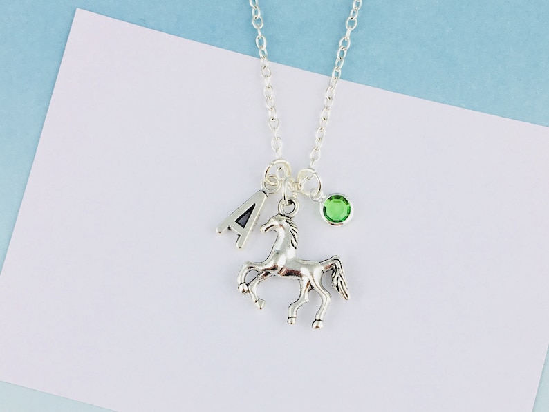 May include: Silver necklace with a horse pendant, a letter "A" charm, and a small green gemstone. The necklace is suspended from a delicate chain. The background is white and light blue.