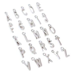 May include: A collection of twenty-six individual silver-toned metal alphabet letter charms, each featuring a small circular loop at the top for attachment. The letters are arranged in a loose grid pattern, representing the complete English alphabet from A to Z.