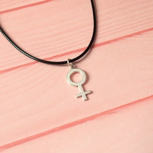 Puede incluir: Un collar con un colgante de plata en forma de s&iacute;mbolo femenino en un cord&oacute;n negro.