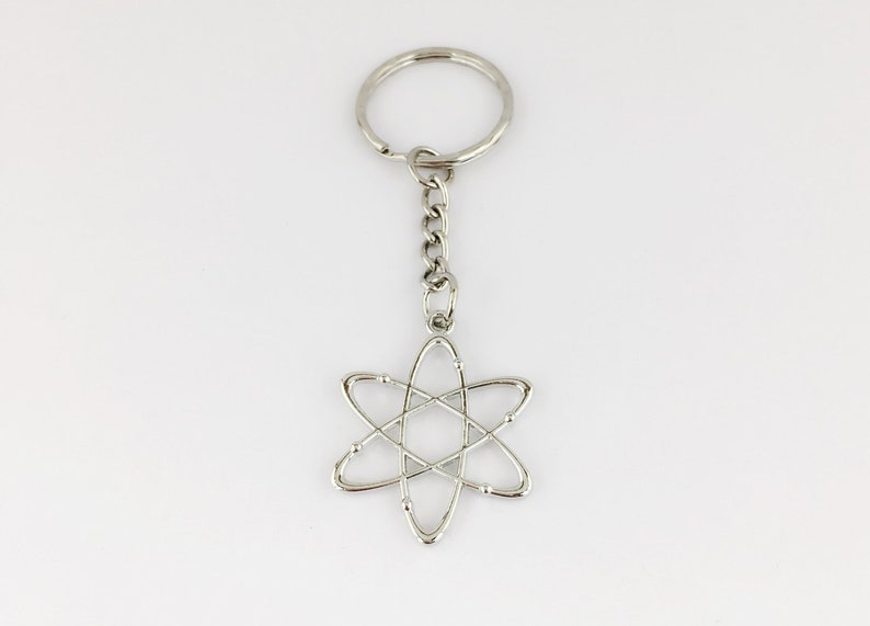 May include: A silver-coloured keyring with a chain and a pendant in the shape of an atom. The atom pendant is a stylized design. The keyring is made of metal and is set against a white background.