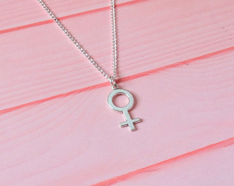 Puede incluir: Un collar de plata con un simple colgante de s&iacute;mbolo femenino sobre un fondo de madera rosa.