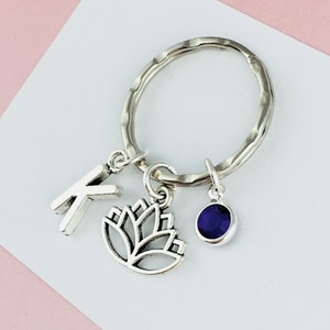 May include: Silver key chain with a letter K charm, a lotus flower charm, and a purple gemstone charm.