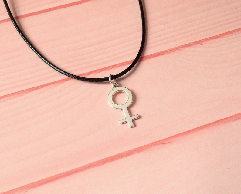 May include: A silver-colored pendant necklace featuring the female symbol. The pendant hangs from a black cord. The background is a light pink wooden surface.