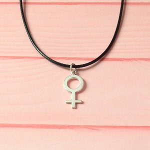 Puede incluir: Un collar de plata con un colgante de s&iacute;mbolo femenino simple en un cord&oacute;n negro.