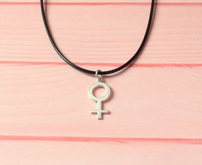 Puede incluir: Un collar de plata con un colgante de s&iacute;mbolo femenino simple en un cord&oacute;n negro.