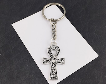 Pode incluir: Chaveiro prateado em forma de ankh. O pingente ankh apresenta detalhes gravados intrincados e é preso a uma corrente e um chaveiro prateado. O fundo é um papel branco sobre uma superfície preta.