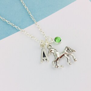 May include: A silver-toned necklace featuring a delicate chain with three dangling charms: a capital letter A, a detailed horse figure in a galloping pose, and a small, round, translucent green gemstone set in a simple silver-toned metal frame.