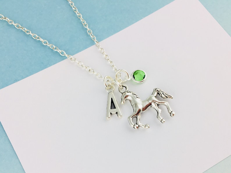 May include: A silver-toned necklace featuring a delicate chain with three dangling charms: a capital letter A, a detailed horse figure in a galloping pose, and a small, round, translucent green gemstone set in a simple silver-toned metal frame.