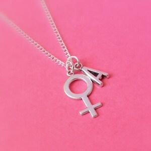 May include: A silver necklace with a charm of the female symbol and the letter 'A'. The necklace is on a pink background.