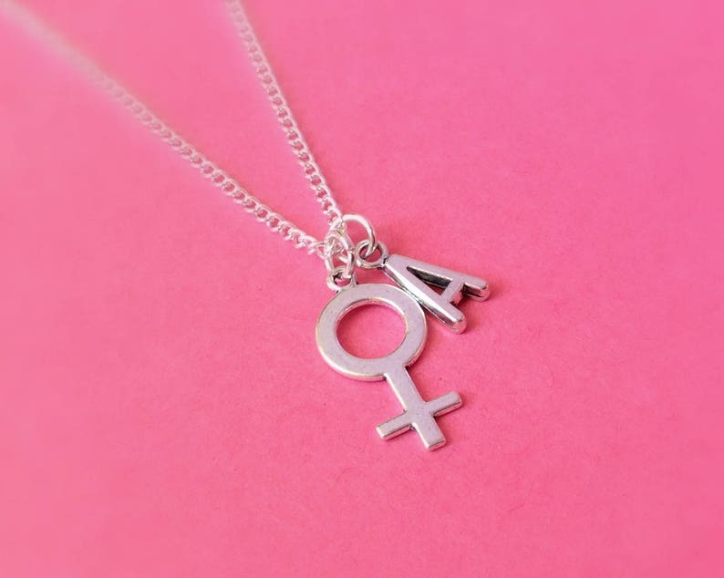 May include: A silver necklace with a charm of the female symbol and the letter 'A'. The necklace is on a pink background.