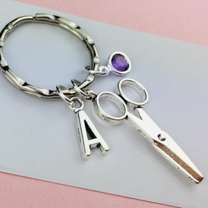 May include: Silver-coloured keyring with a pair of scissors charm, a letter "A" charm, and a purple gemstone accent. The keyring has a textured ring and is displayed on a white surface with a pink background.